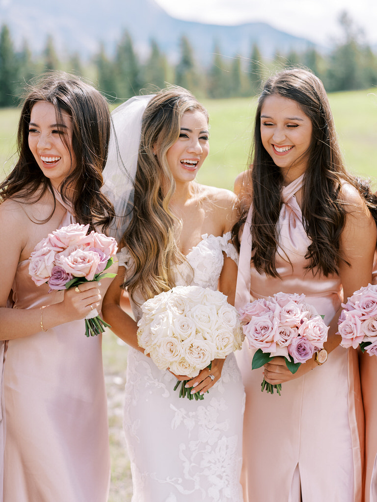 candid wedding party portraits in Canadian Rockies. Bride  with  bridesmaids. Pink satin dresses with pink and white roses. 