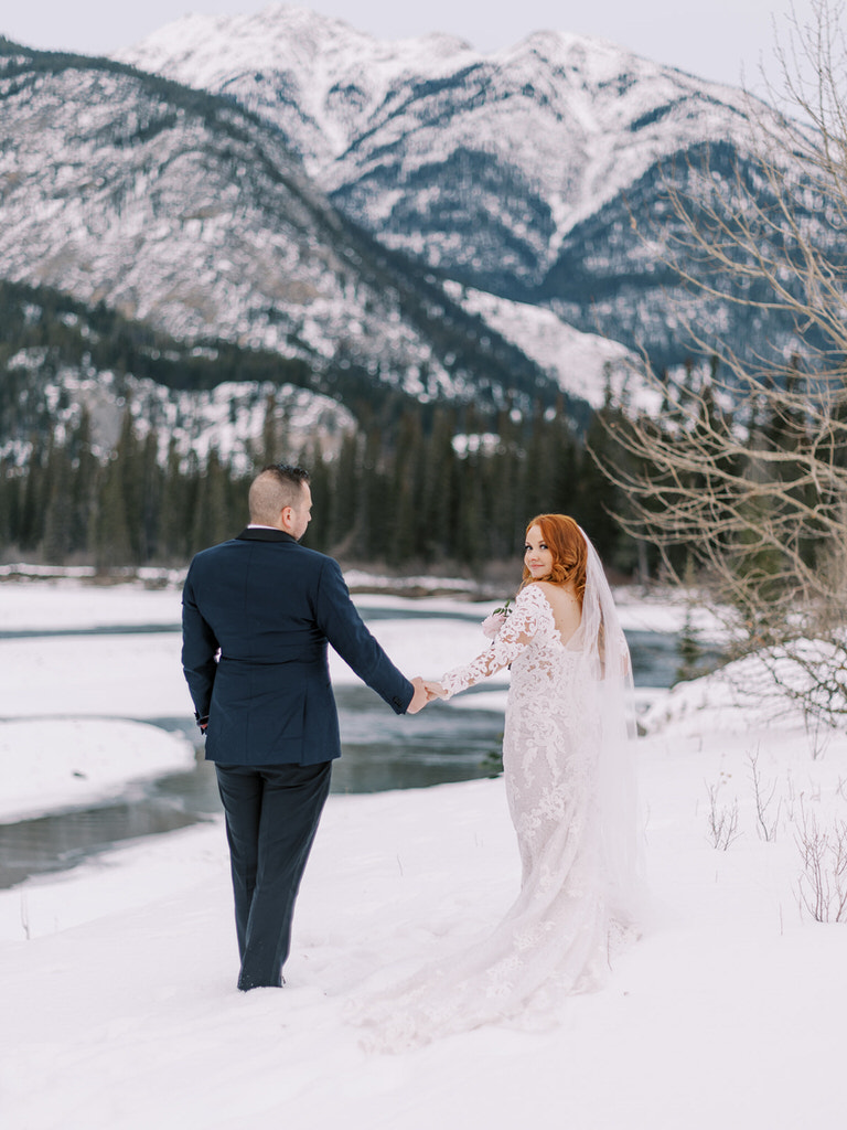 Bride and Groom wedding portrait shows couple walking along river, Bride is looking back over should with winter mountain landscape, Banff, Canada  