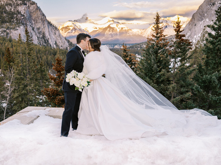 Mountain bride in strapless wedding dress with veil and fur cape, couple portrait bride and groom kiss with mountains in the background.  Fairmont Banff Springs in Banff, Canada. 