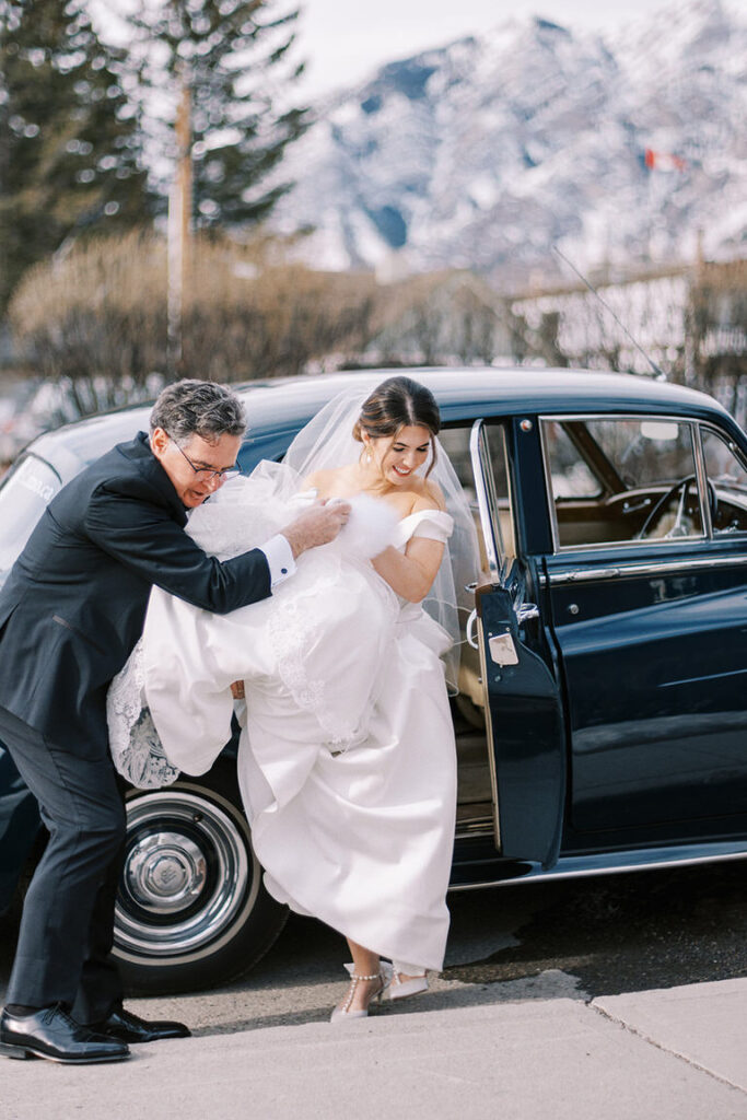 Mountain bride in strapless wedding dress, with veil exits car to wedding ceremony with father of bride in Banff, Canada. 