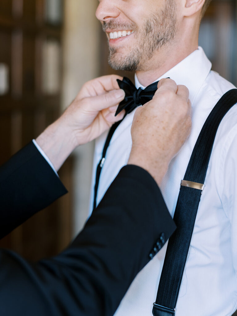 Groom getting ready, black tuxedo, tying a bowtie. wedding photography