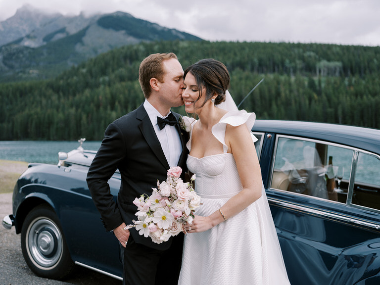 bride and groom couples portrait, with vintage car and mountain background, off shoulder dress, and wedding florals of light pink and white. 
