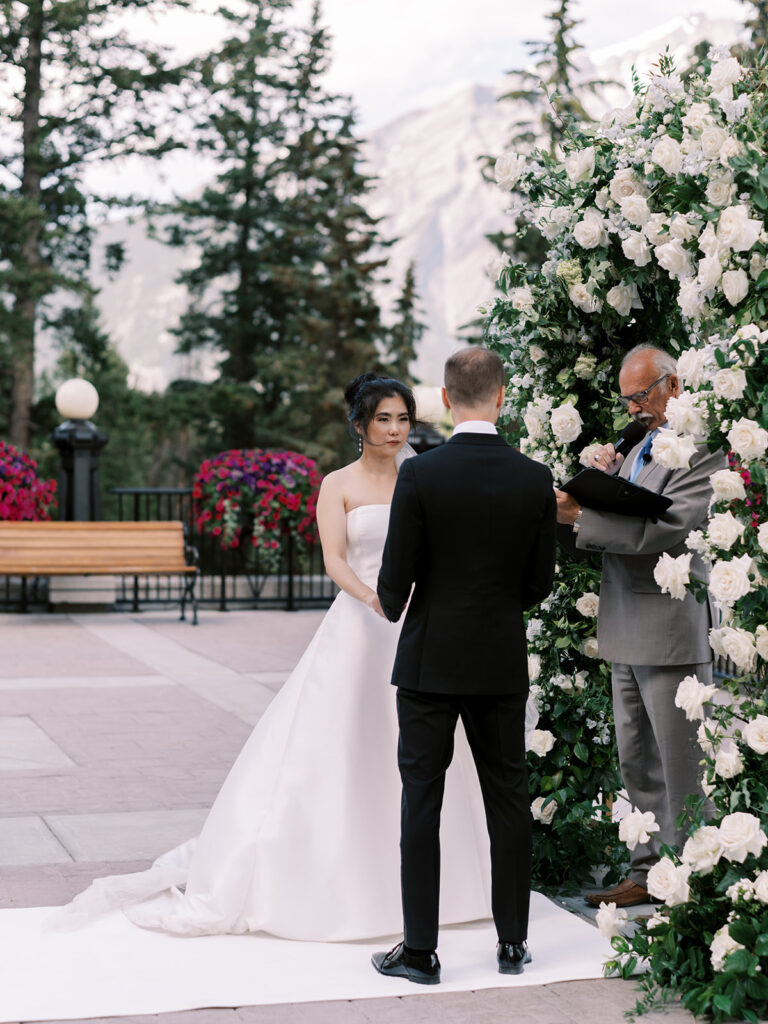 Bride and groom exchanging vows. Wedding ceremony, outdoor wedding with white florals and green arches. Banff Springs Fairmont Hotel 