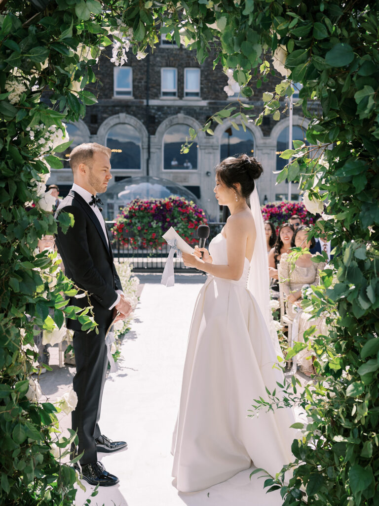 Bride and groom exchanging vows from behind the alter. Wedding ceremony, outdoor wedding with white florals and green arches. Banff Springs Fairmont Hotel 