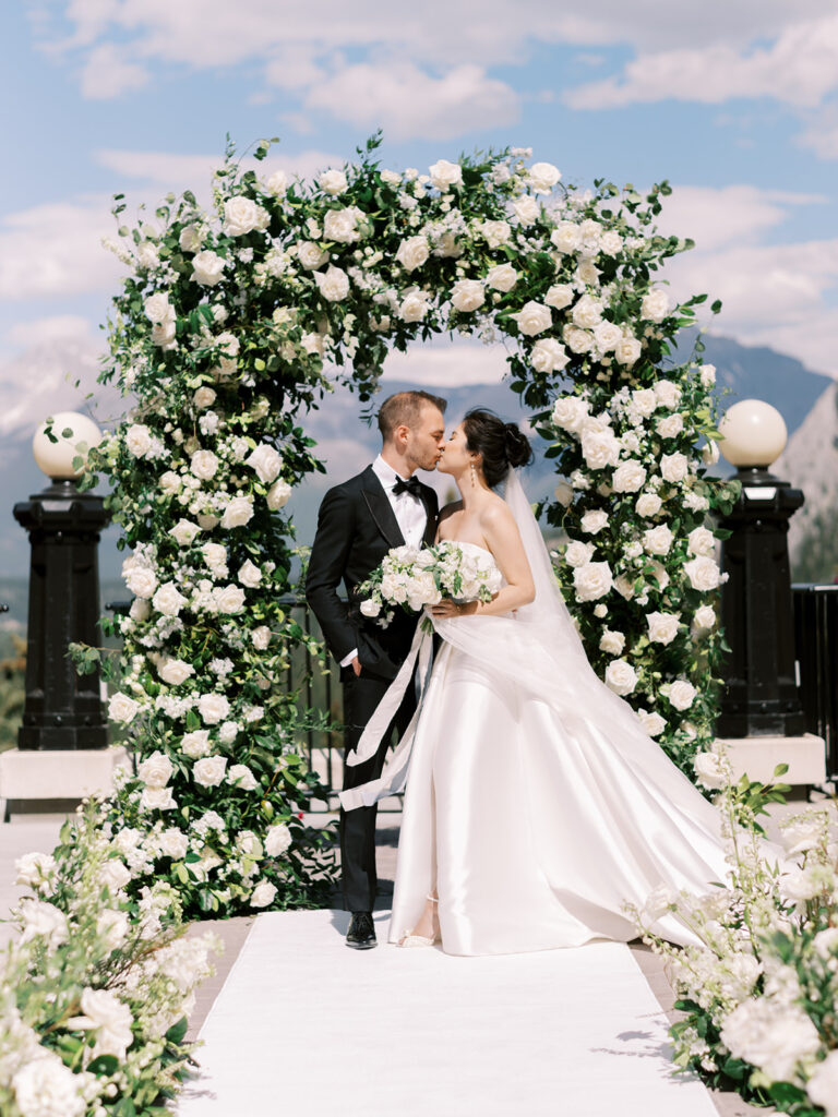 Bride and groom first kiss. Wedding ceremony, outdoor wedding with white florals and green arches. Banff Springs Fairmont Hotel 