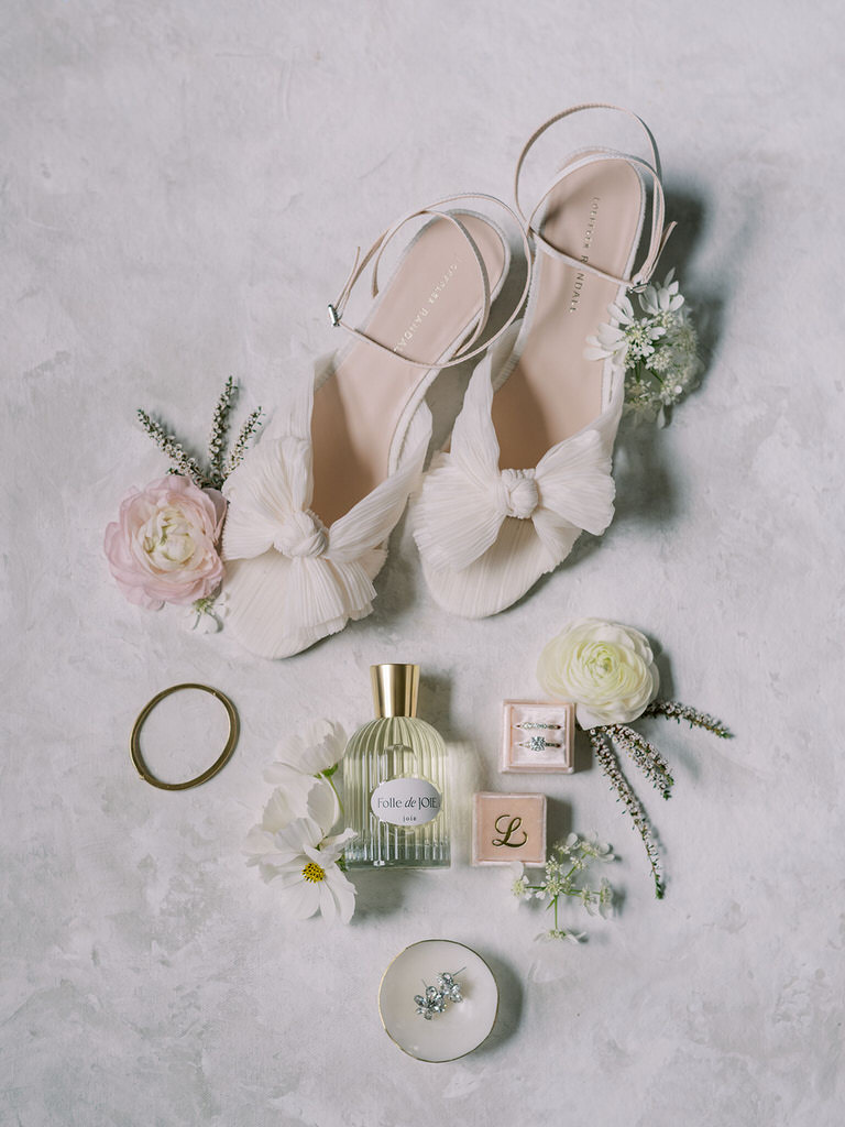 Bridal flatlay with wedding details, including perfume, blush pink and white florals, gold bracelet and diamond earrings. Personalized ring box with bride's initials