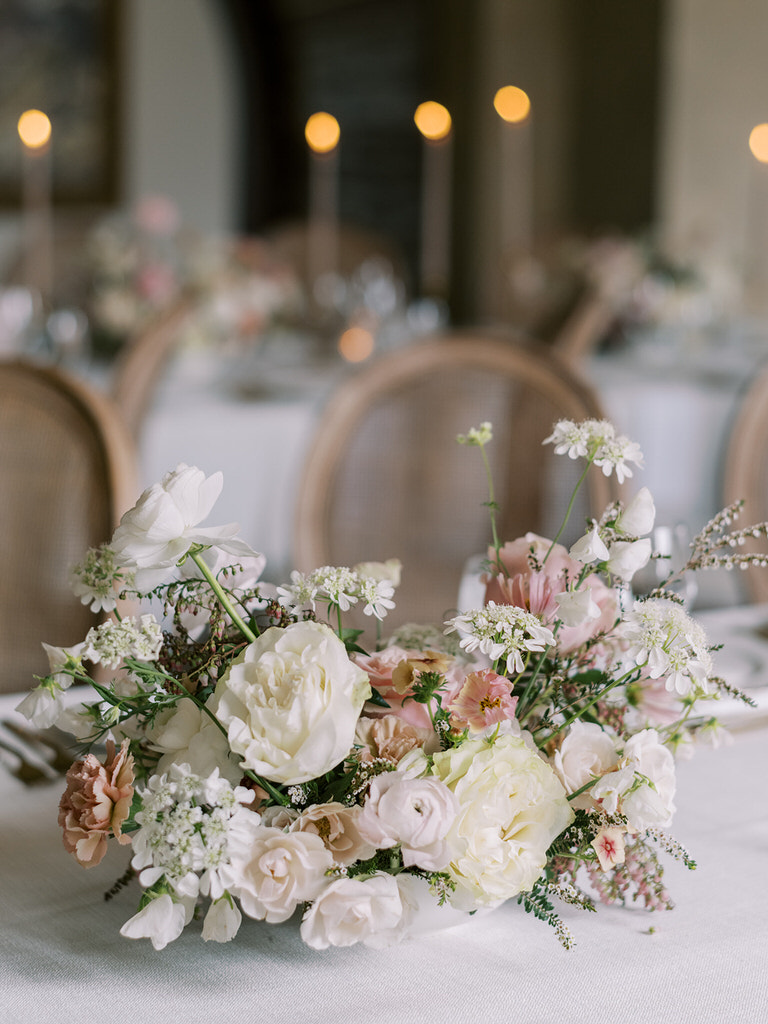 wedding reception space in Fairmont Banff Springs hotel. Light pink pastel and white florals, gold features, white linens, and long candles accent wedding table. Beige rattan chairs. 