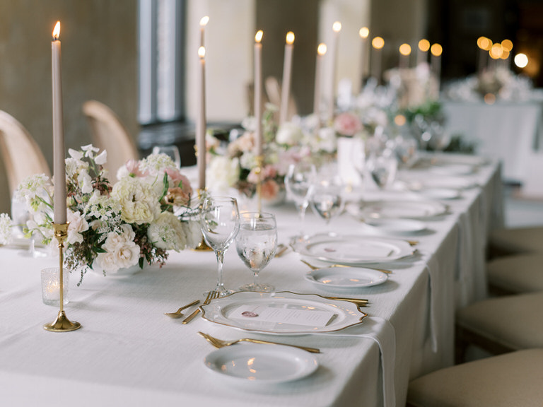wedding reception space in Fairmont Banff Springs hotel. Light pink pastel and white florals, gold features, white linens, and long candles accent wedding table. Beige rattan chairs. 