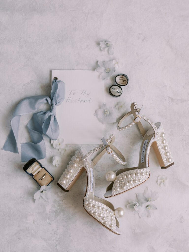 wedding flatlay of bride's vows with light blue ribbons, white florals, pearl jimmy choo wedding shoes, wedding rings, and white airy vibe. 