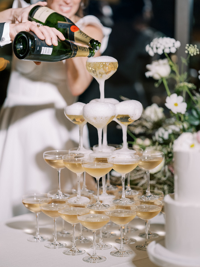 bride and groom pouring champagne at top of champagne tower. candid wedding shot. Wedding reception space in Fairmont Banff Springs hotel. Light pink pastel and white florals, gold features, white linens. 