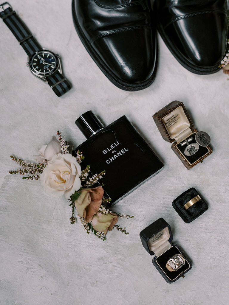 Groom flatlay with wedding details. Color scheme of black, blush pink florals. Wedding details of cologne, boutonnière, cufflinks, watch and wedding band.  