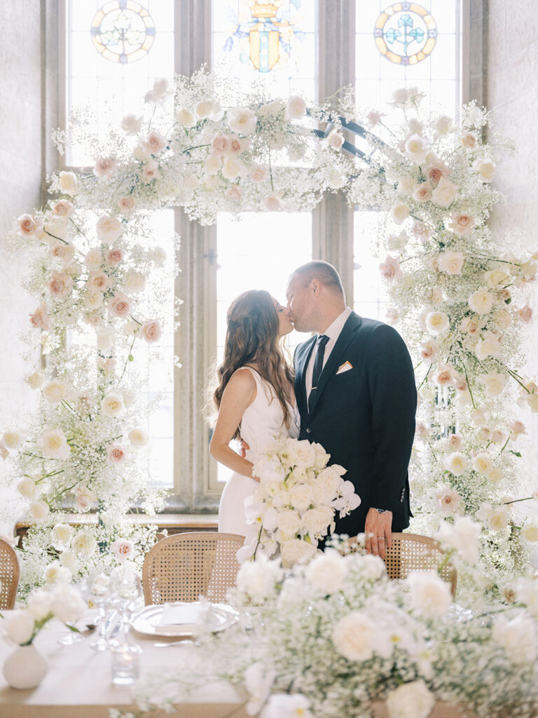 Banff springs wedding photographer captures couple kissing in front of modern floral arch covered in roses and babys breath in the stunning Mt. Stephen Hall at the Fairmont Banff Springs Hotel