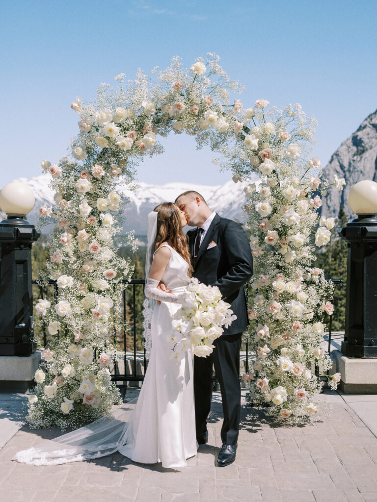 Banff springs wedding photographer captures couple kissing in front of modern floral arch covered in roses and babys breath on the Terrace of the Fairmont Banff Springs Hotel