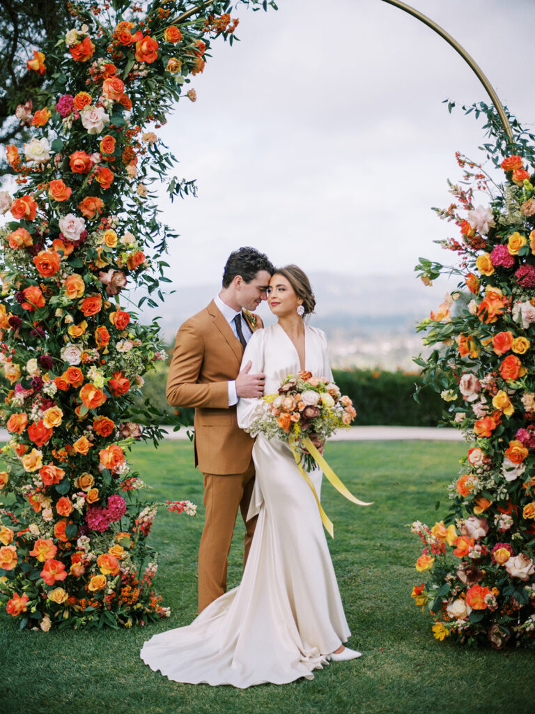 Bride and groom standing under a bright full floral arch at California Wedding