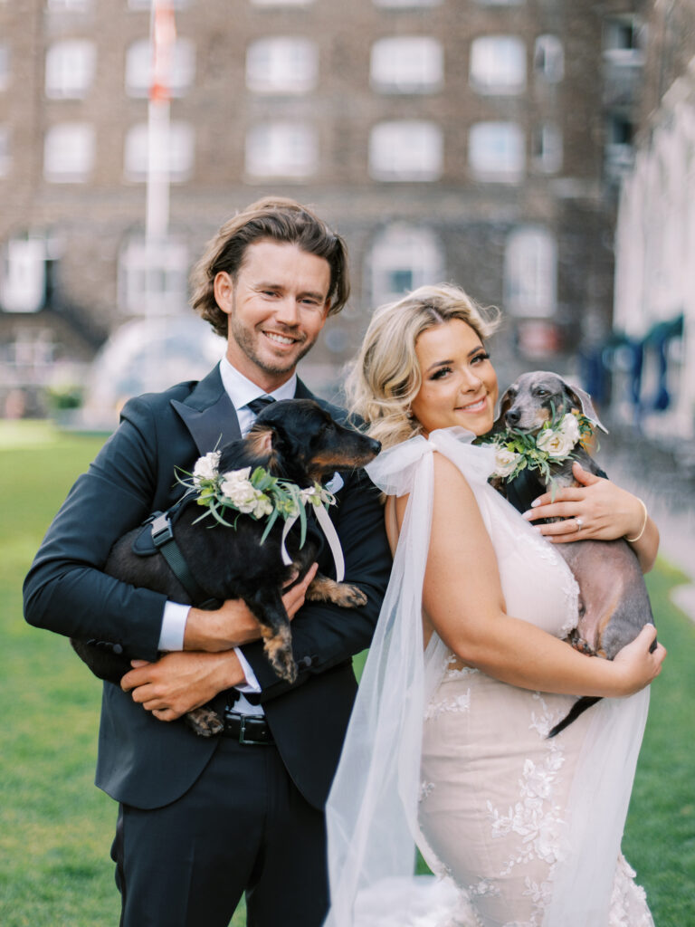 bride and groom with dogs with floral collars on, posing in front of the Fairmont Banff Springs Hotel. captured by Banff Wedding Photographer Justine Milton
