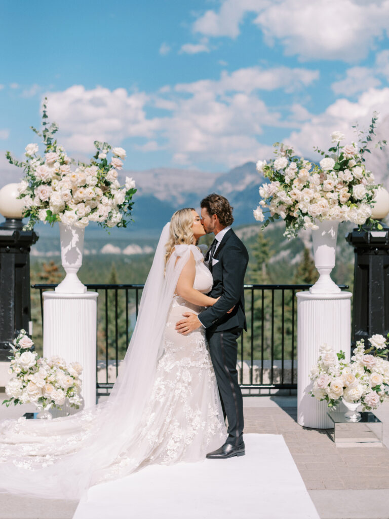 Fairmont Banff Springs Hotel wedding ceremony on the terrace. Ceremony decor with white and pink florals in overflowing urns on ribbed pillars. Bride in a wedding dress with tulle bows captured by Banff Wedding Photographer Justine Milton