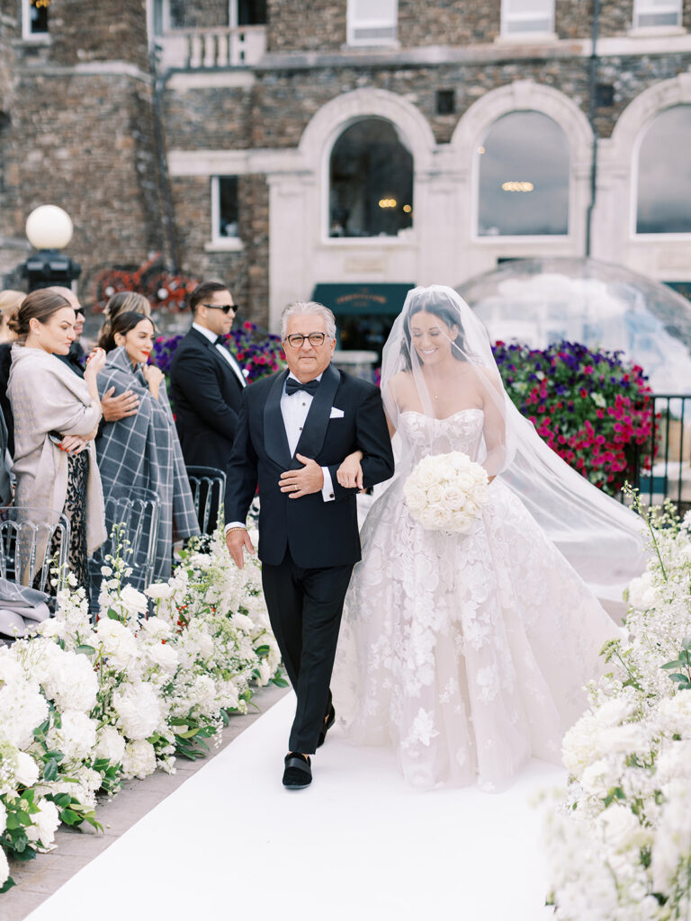 bride and dad walk down an elegant ceremony aisle filled with lush white florals at the Banff Springs hotel terrace