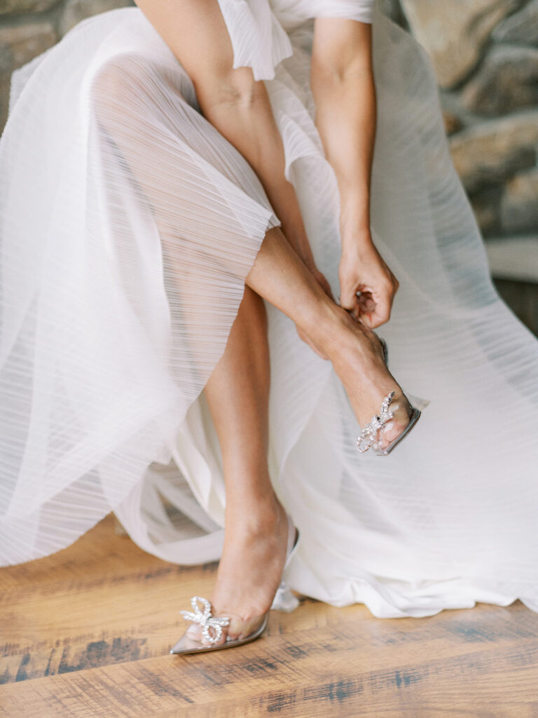 bride getting ready putting on clear shoes with silver bow captured by Canadian and destination wedding photographer Justine Milton